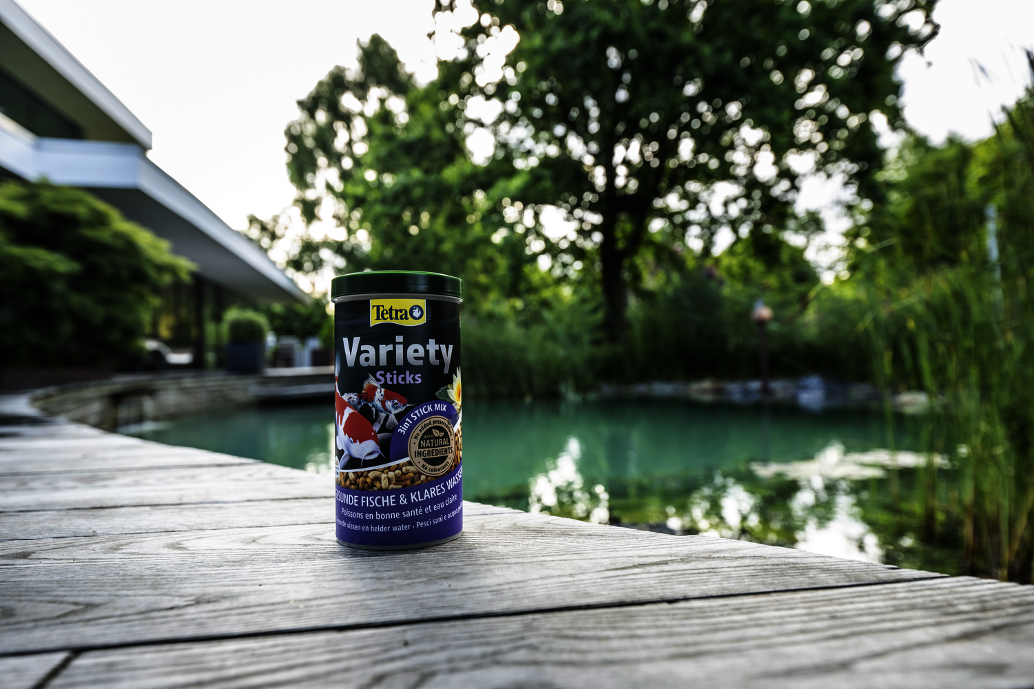tetra-pond-variety-sticks-havuz-balik-yemi-1-litre-5 Bahçe havuzu kenarında ahşap zemin üzerinde duran Tetra Pond Variety Sticks balık yemi kutusu